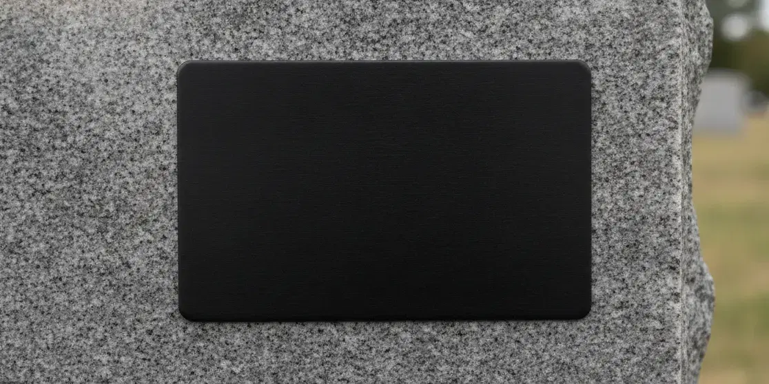 A black metal plaque the size of a credit card fixed to the face of a grey granite headstone